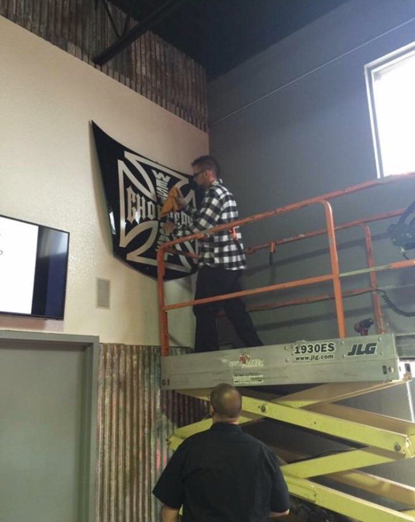 owner travis hanging a west coast choppers sign on wall with scissor lift