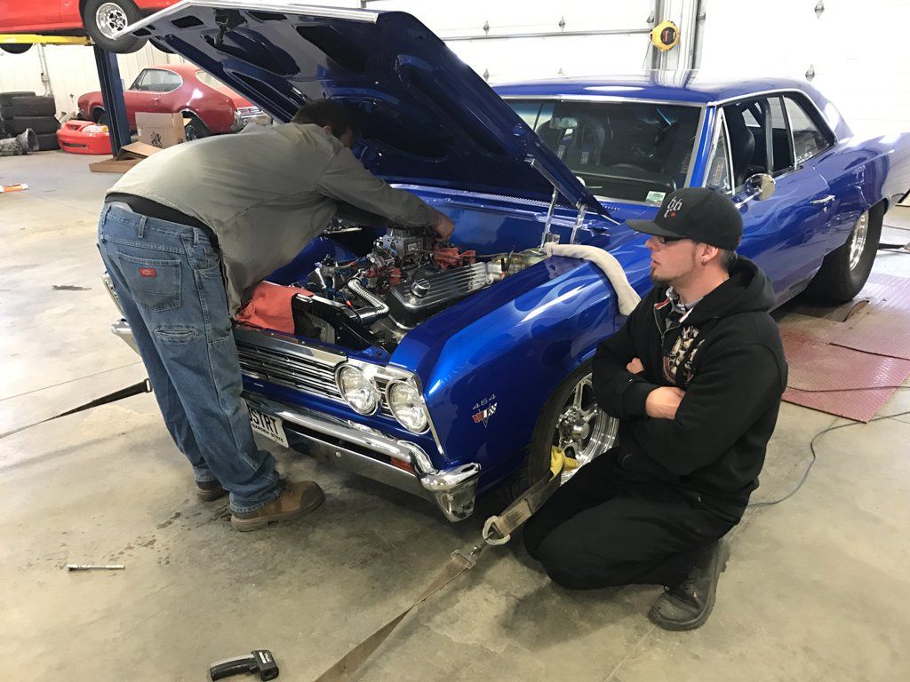 one mechanic working on car, one mechanic sitting next to car with hood open