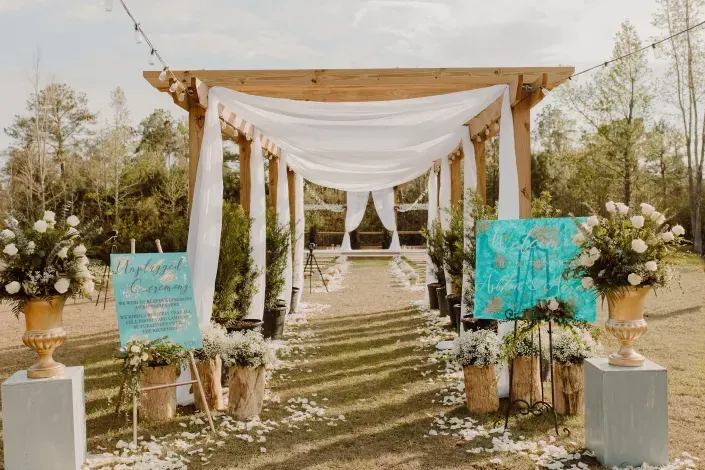 Outdoor wedding aisle with white drapery, wooden arch, floral arrangements, and turquoise signs along a stone path