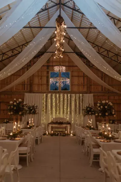 Decorated barn wedding reception with long tables, white drapery, and warm chandelier lighting