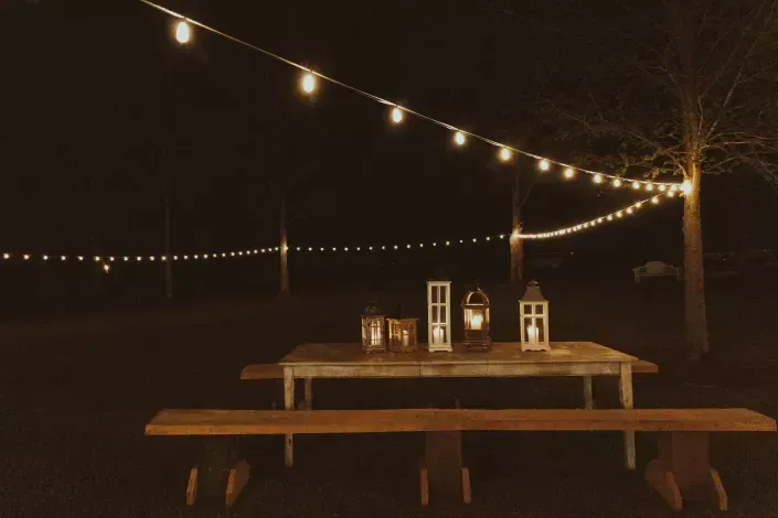 String-lit outdoor picnic table at night with lanterns and a tree in the background