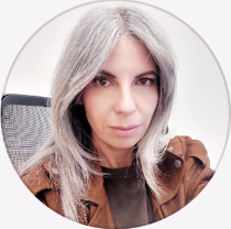 Mujer con cabello largo y gris con una chaqueta de gamuza marrón, mirando directamente a la cámara.