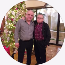 Dos hombres posan del brazo frente a un árbol de Navidad. Uno lleva camisa abotonada y pantalón negro, el otro, camisa a cuadros y chaqueta.