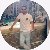 Hombre con camiseta blanca y pantalones claros, apoyado en una repisa de piedra cerca de una fuente.