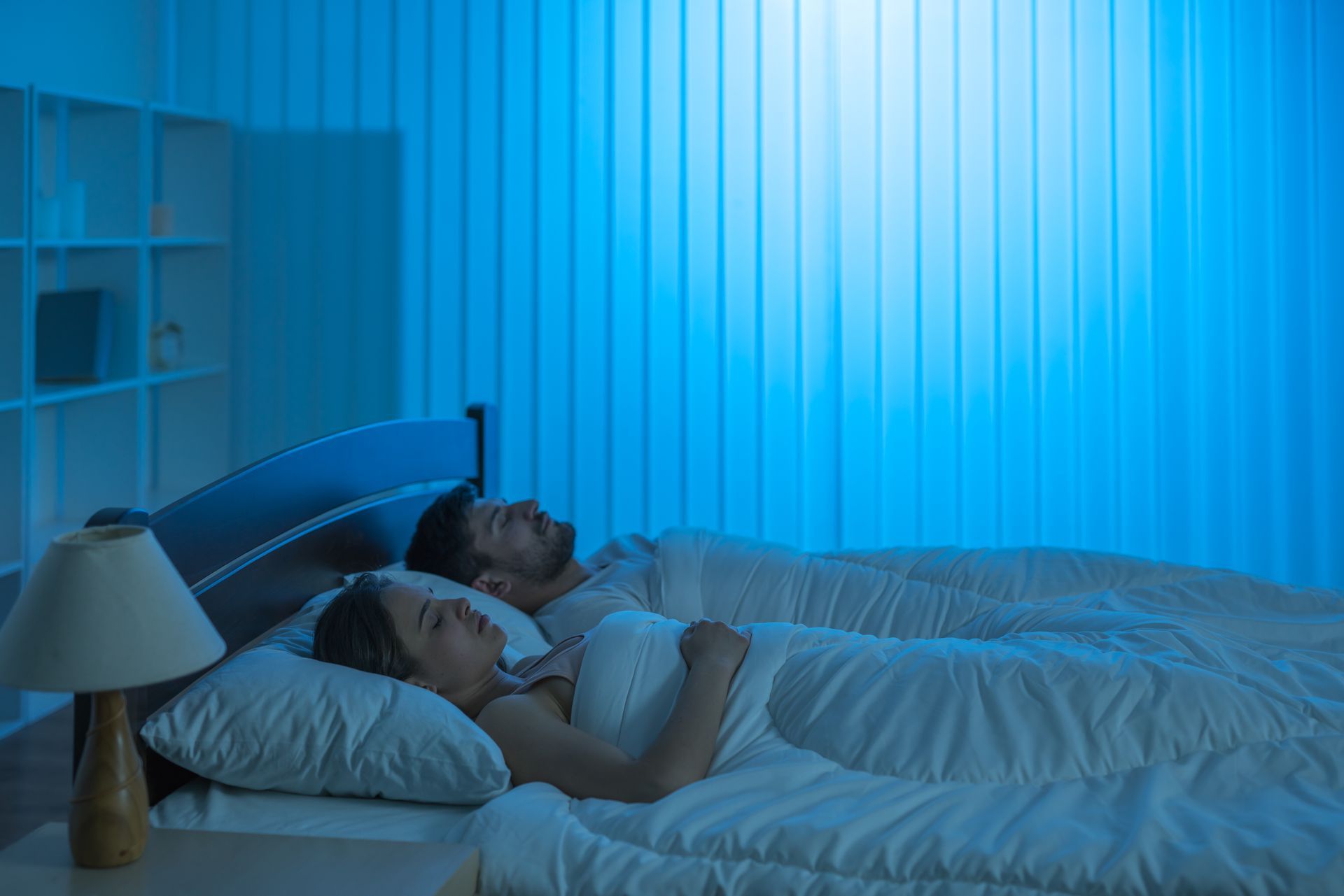 A man and a woman are sleeping in a bed with blue blinds.