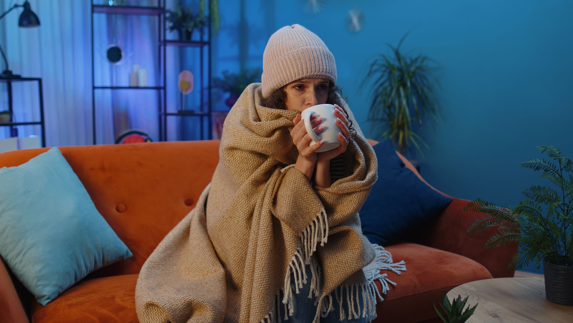 A woman wrapped in a blanket is sitting on a couch drinking a cup of coffee.