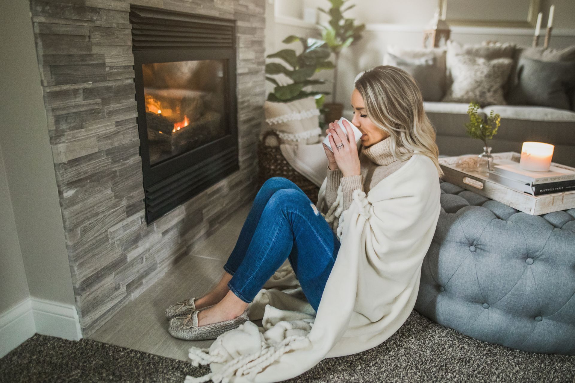 A woman is sitting on the floor in front of a fireplace drinking coffee.