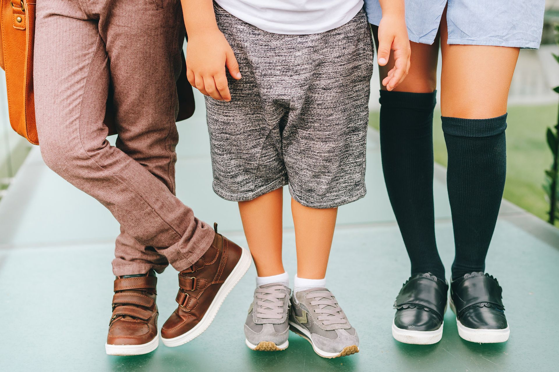 Three people standing close together; one in brown pants and shoes, one in shorts and sneakers, and one in a dress with black socks and shoes.