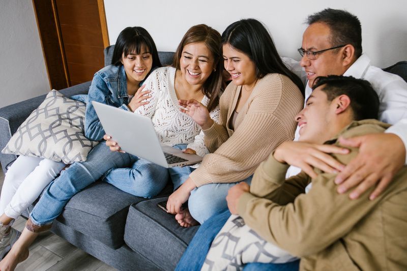 Group of five people sitting on a couch, smiling and looking at a laptop together in a living room.