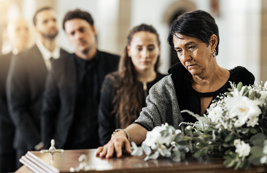 Mourners stand by a casket with white flowers, one person leaning forward in grief.