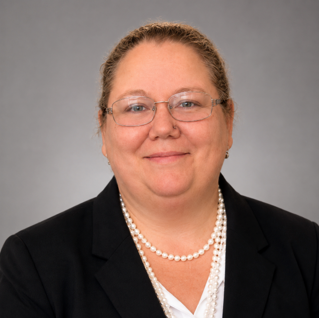 Studio headshot of a smiling person in glasses, black blazer, white shirt, and pearl necklace.