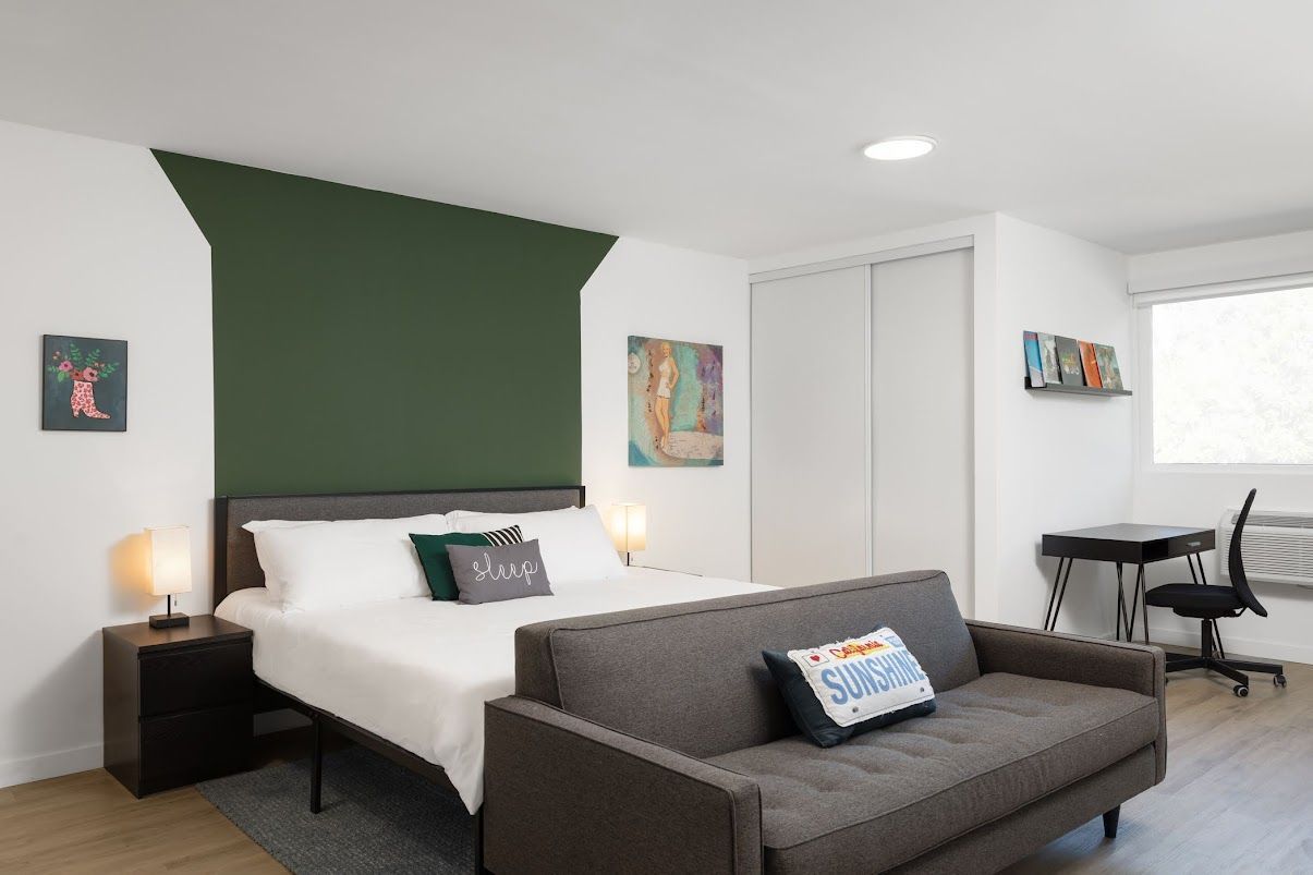 Photo showing a room with a bed against a wall and a desk area in the background