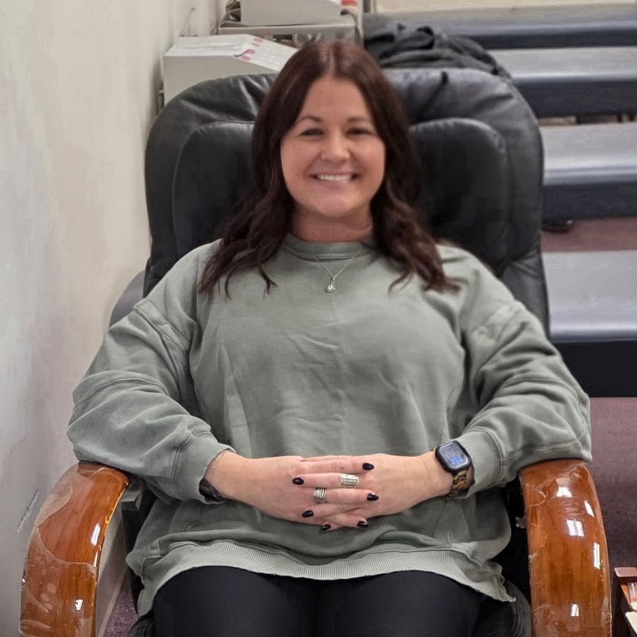 Woman in a green sweater, smiling, sitting in a nail salon chair, hands crossed, dark nail polish.