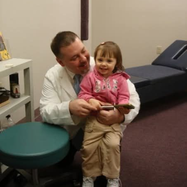 Doctor and young child smiling in an office setting. The doctor holds a small item, child sits on his lap.