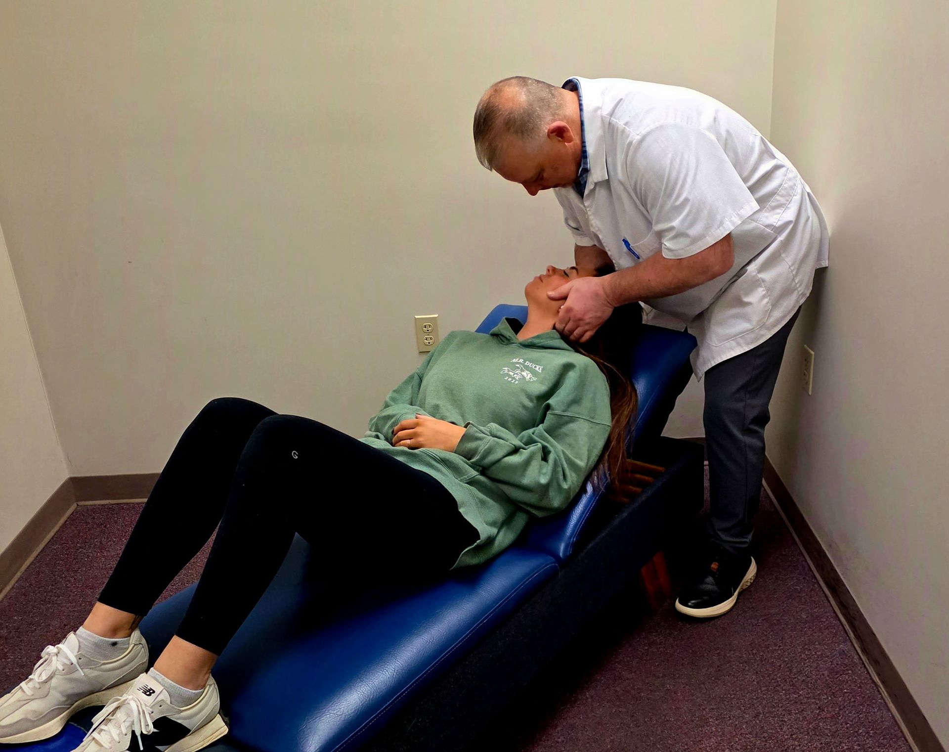Doctor Joe examining patient's neck. Patient reclines on blue exam table in room with purple carpet.