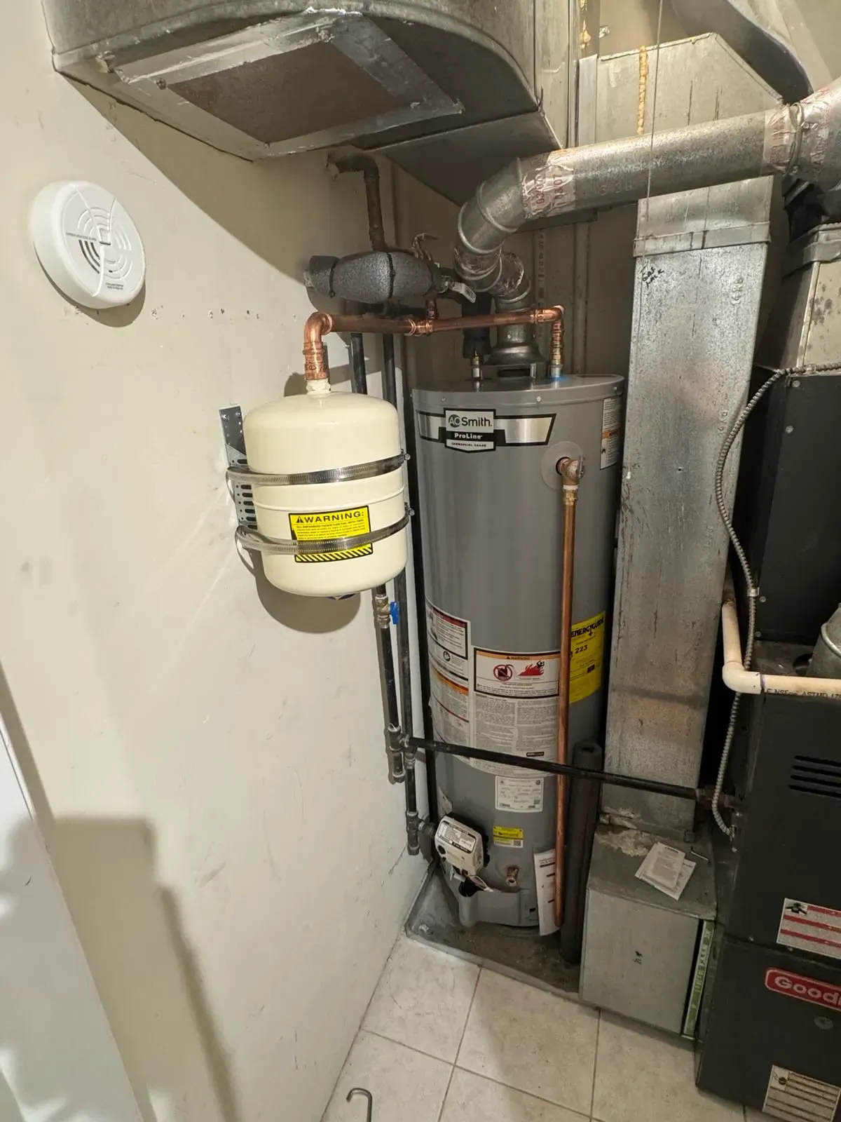 Water heater and expansion tank in a utility room. Copper pipes, metal ductwork, and white walls.