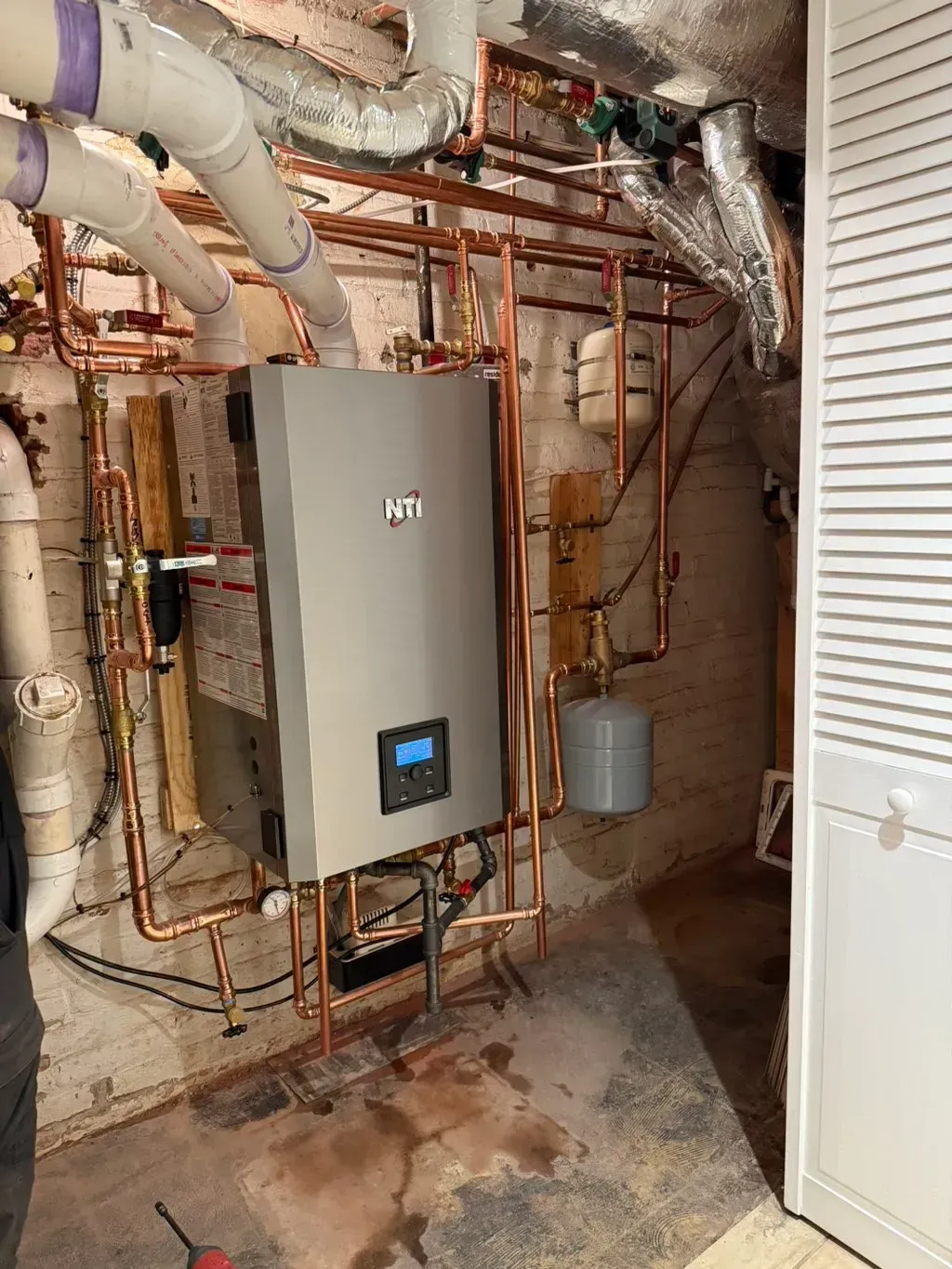 Copper pipes and a gray boiler installed in a basement, with a white door to the right.