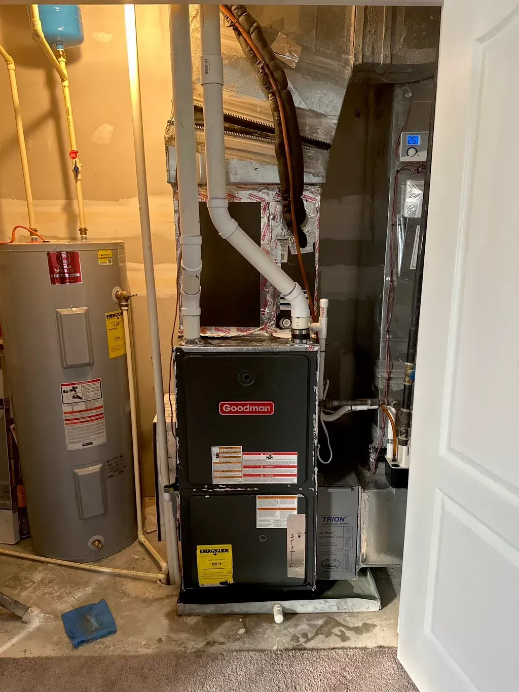 A furnace and water heater are in a basement. The Goodman furnace has a black door. Plumbing and ductwork are visible.