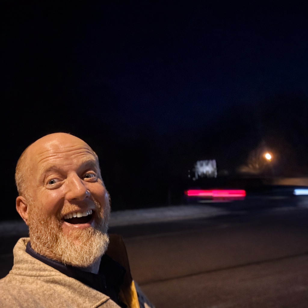 Man with beard smiling widely at night, blurred lights in the background.