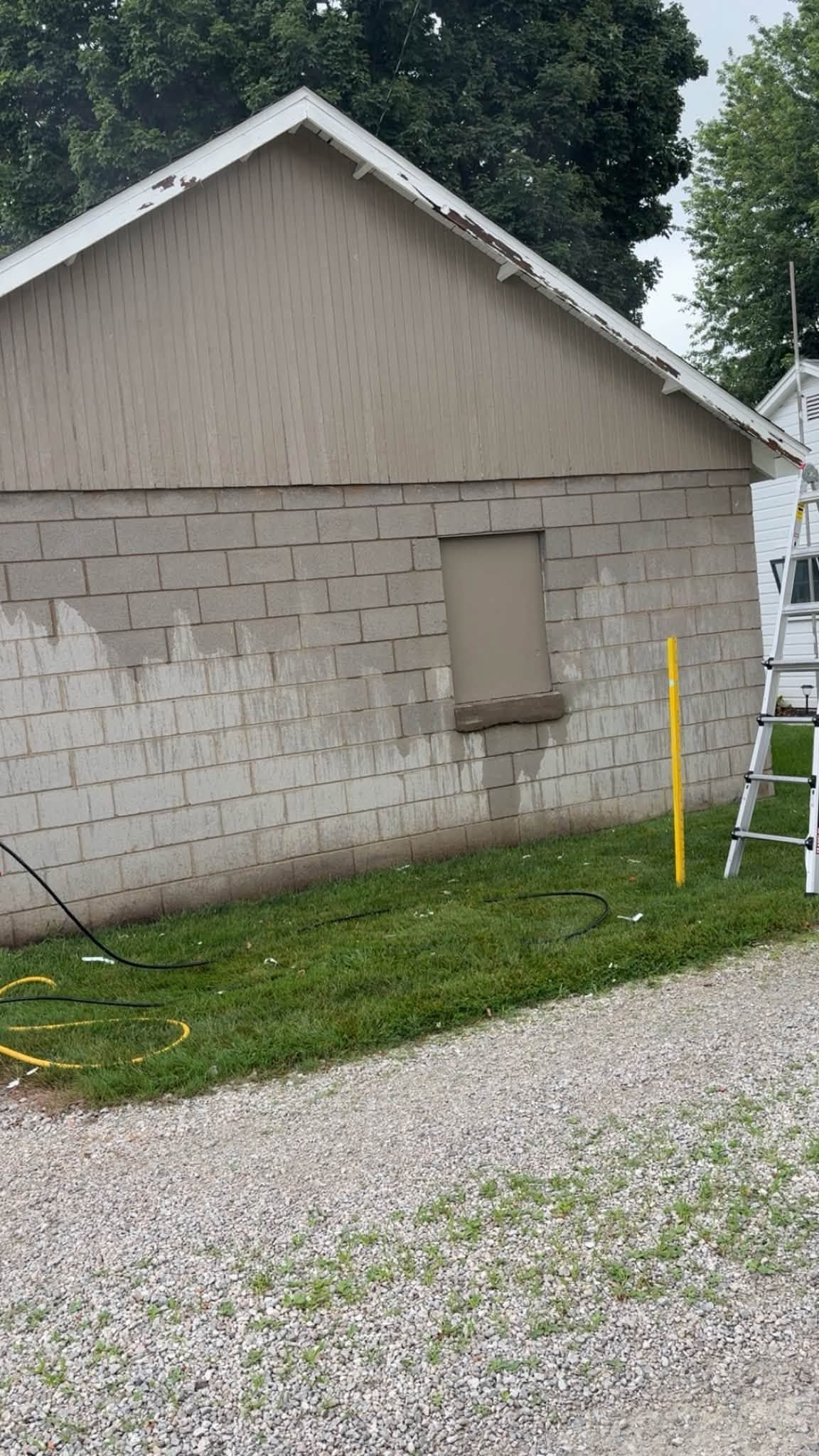 Side of beige house with boarded window, gravel driveway, grass, and ladder at right edge