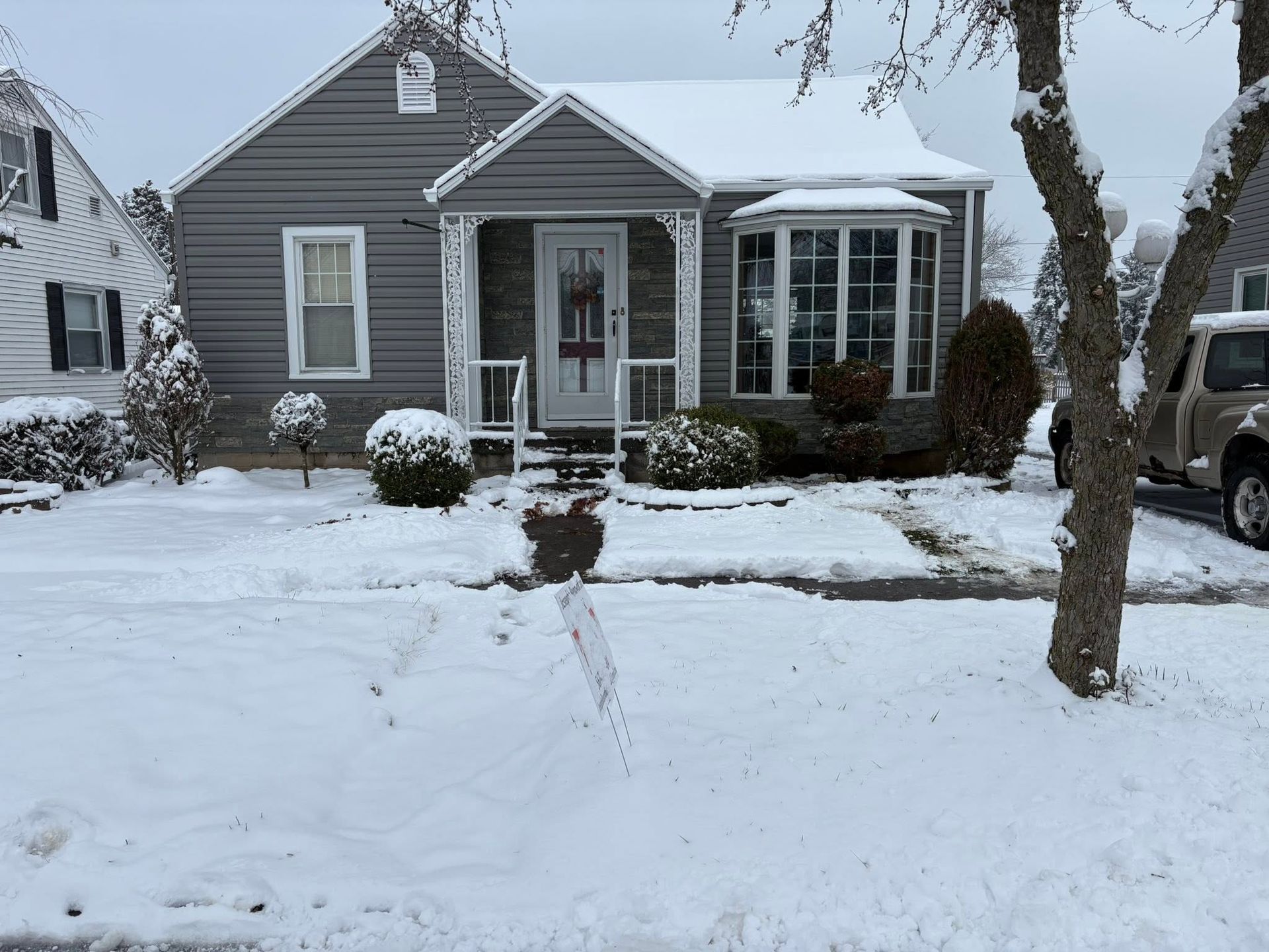 Snow-covered gray house with white trim and a bay window, fronted by a snowy yard and bare trees.