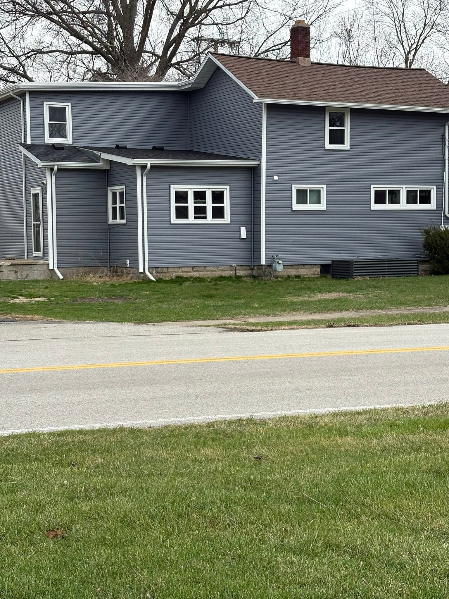 Gray two-story house with white trim and a red brick chimney beside a road.