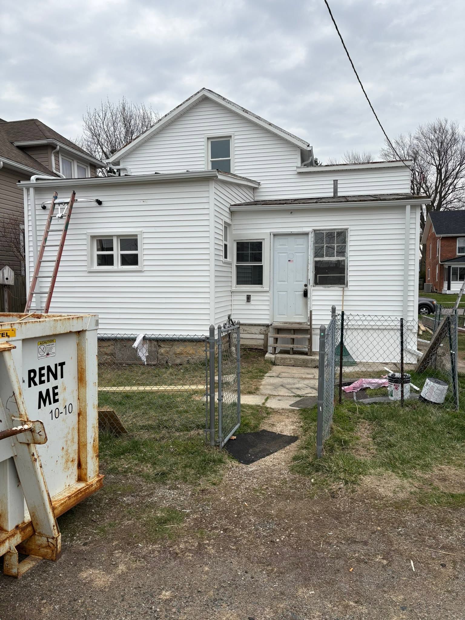 Small white house with boarded front door and a “RENT ME” dumpster in the yard