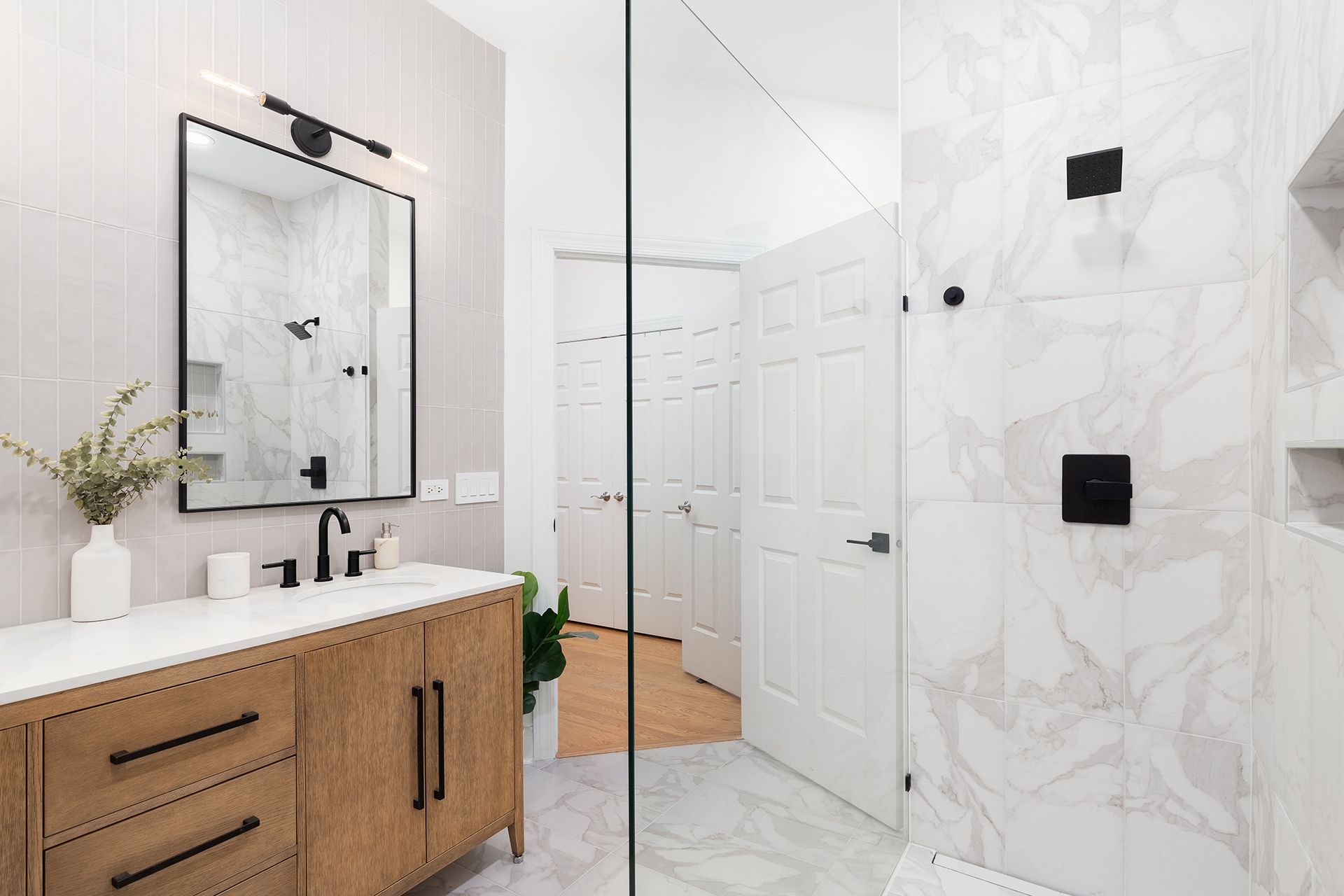 Bright bathroom with wood vanity, large mirror, glass shower, white marble tile, and black fixtures