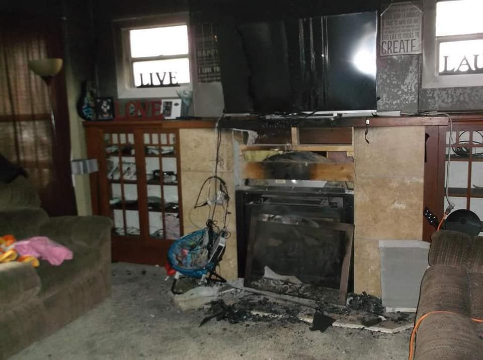 Living room damaged by fire, charred fireplace, blackened walls, TV, and debris on the floor.