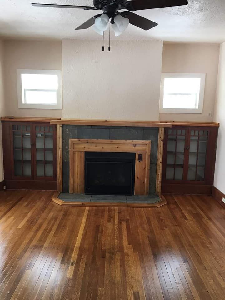 Living room with a fireplace, built-in cabinets, wood floors, and two windows.