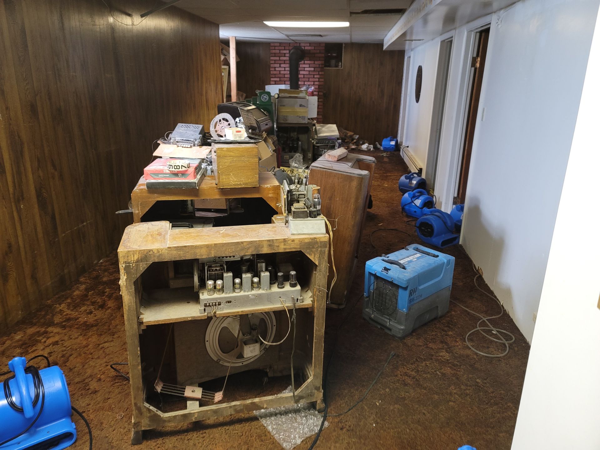 Flooded basement with old electronics, two dehumidifiers, and water damage on wood-paneled walls.