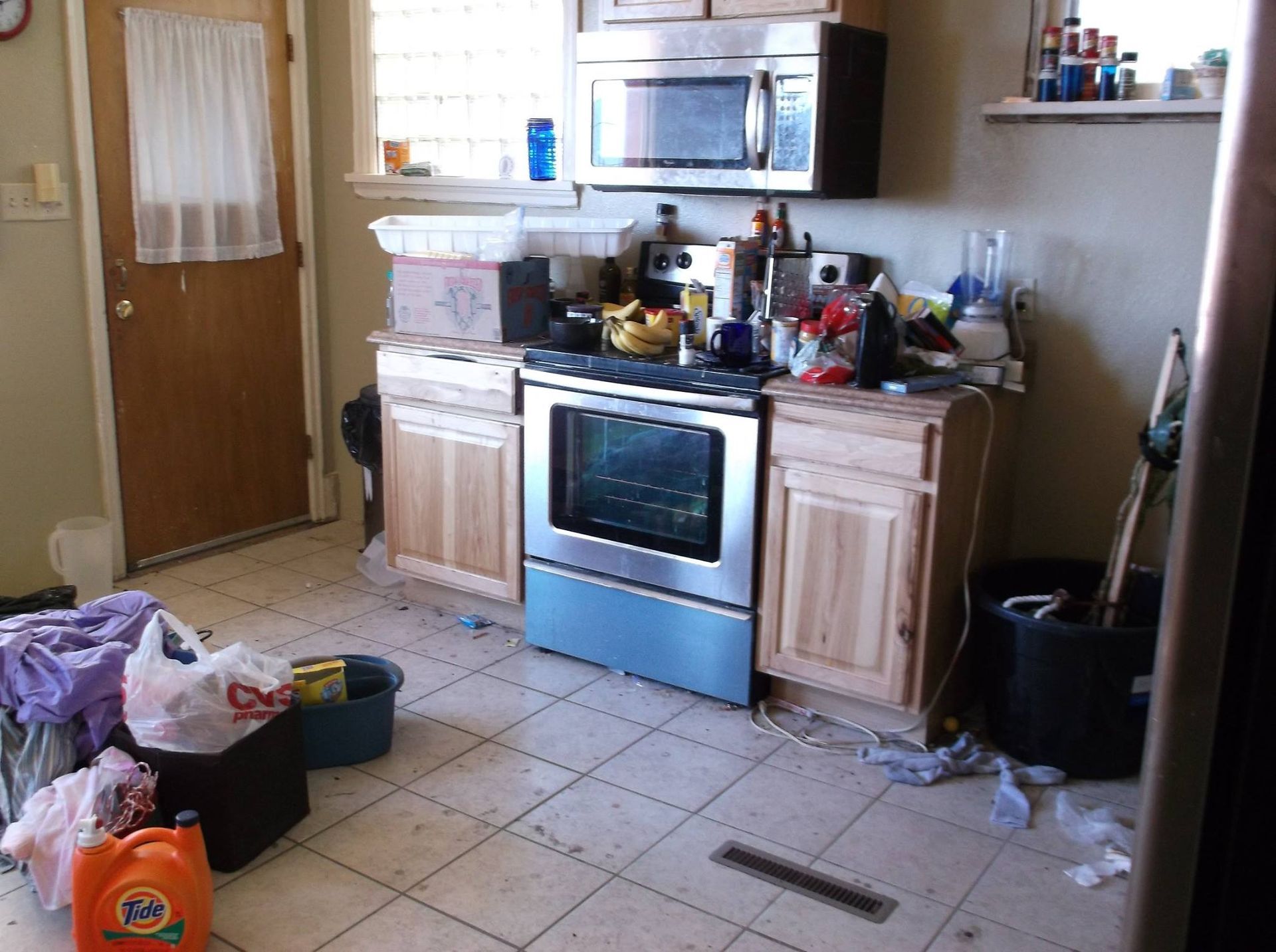 Cluttered kitchen with stained tile floor. Stove, microwave, cabinets, and various items piled around.