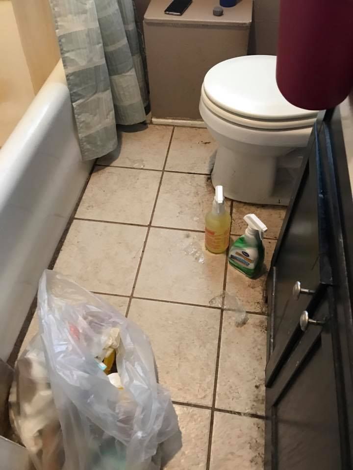Bathroom with a dirty tile floor, bathtub, toilet, trash bag, and cleaning supplies.