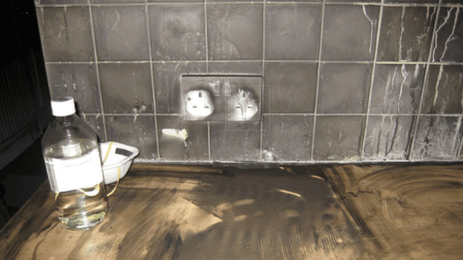 Burned kitchen: dark countertop, wall tiles, damaged electrical outlet, bottle, and small container.