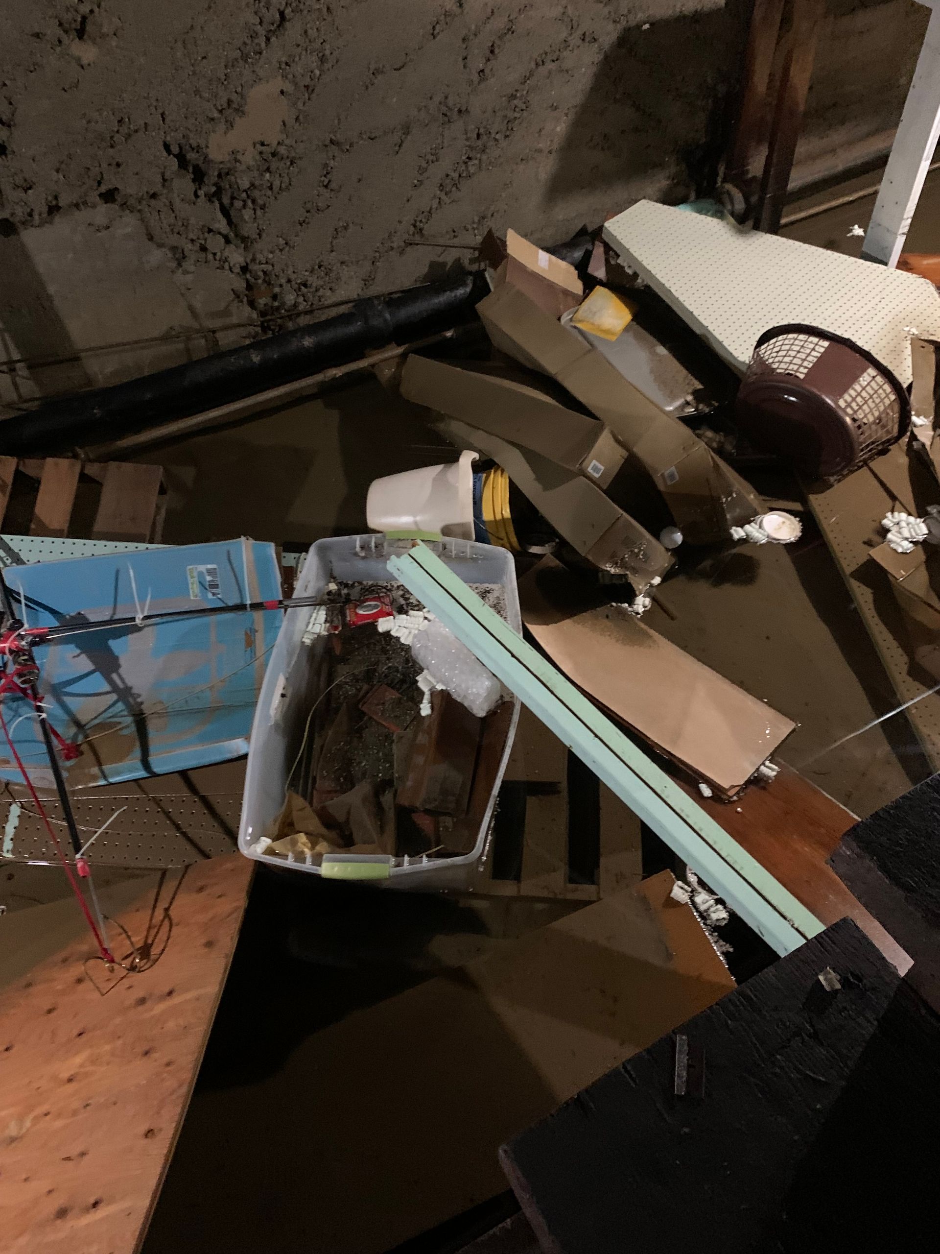 Overflowing storage area: cardboard, blue bin, furniture, and debris piled in a basement corner.