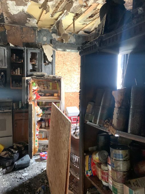 Kitchen interior heavily damaged by fire. Burnt ceiling, debris, open refrigerator, charred shelves with cans.
