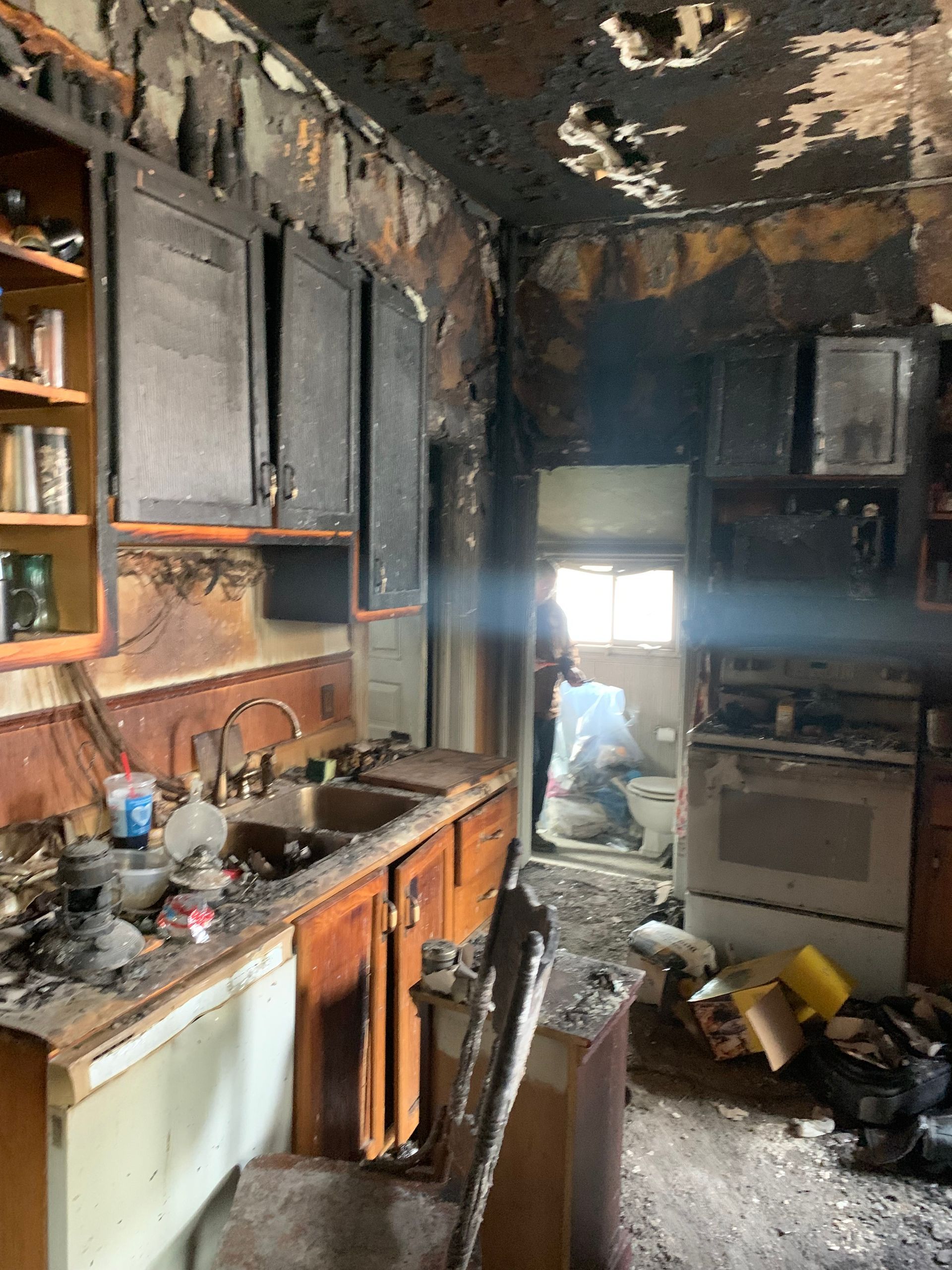 Fire-damaged kitchen interior, walls and cabinets charred black. Debris scattered, person visible in doorway.