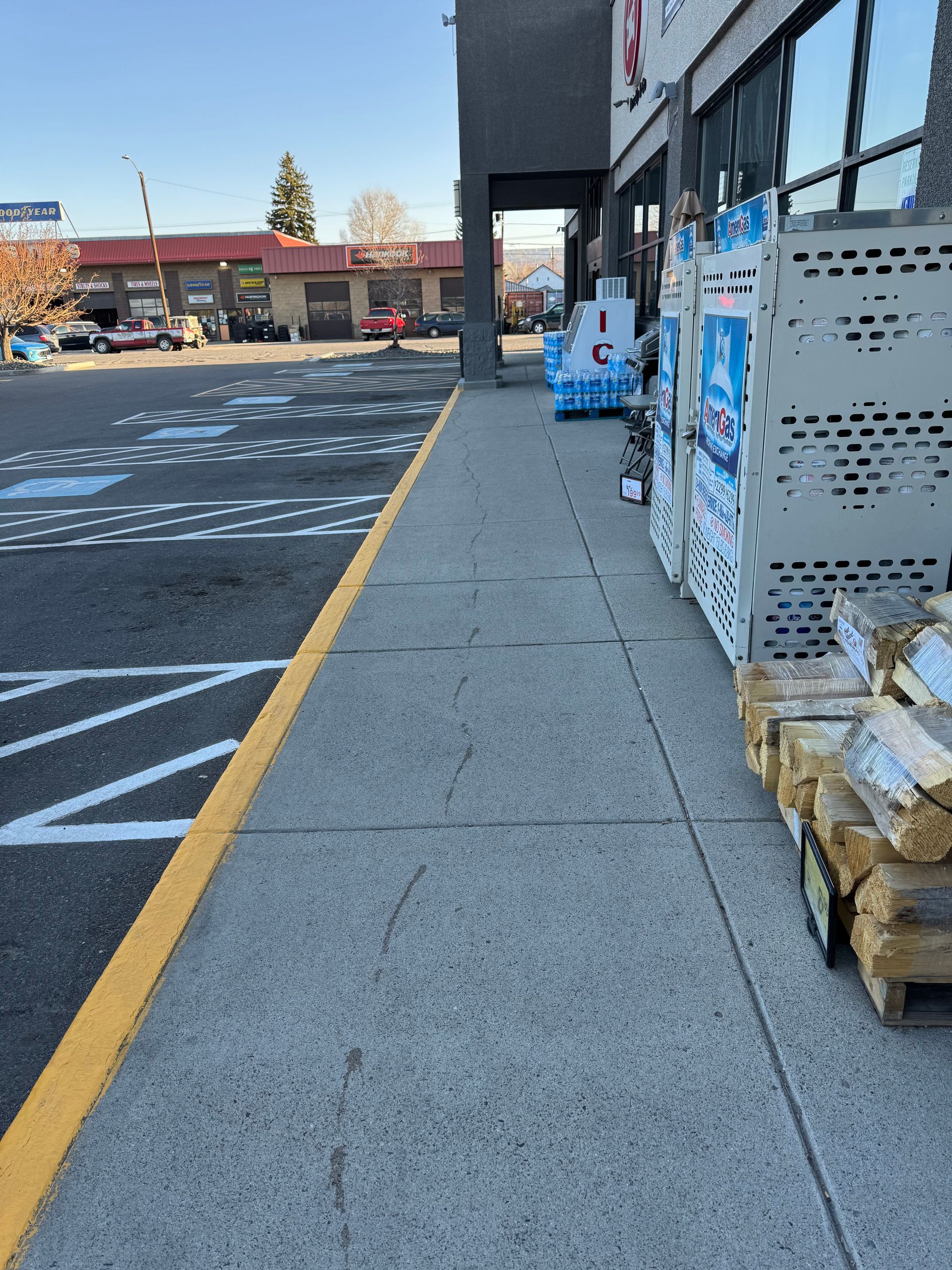 Sidewalk next to a parking lot and store with firewood and bottled water displayed.