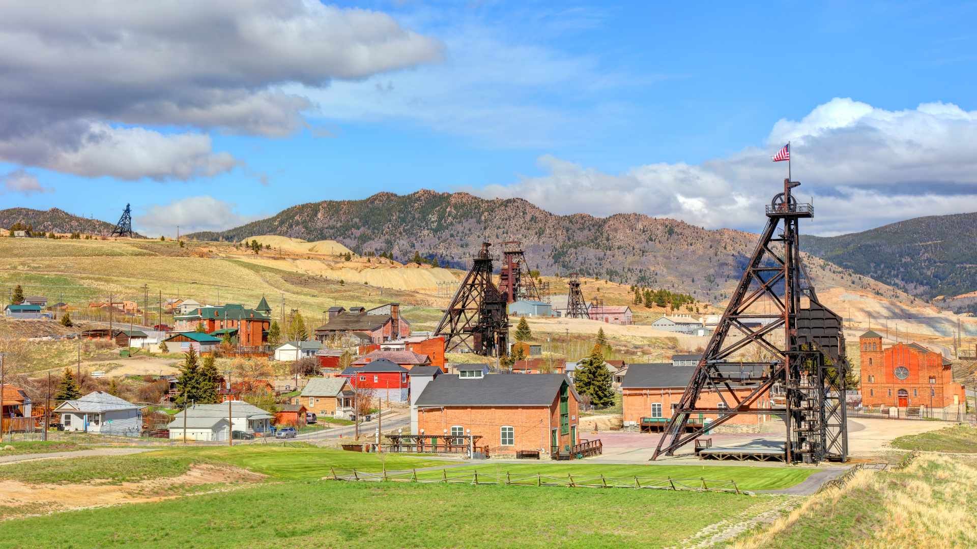 Historic mining town with wooden headframes, red brick buildings, and rolling hills under a blue sky.
