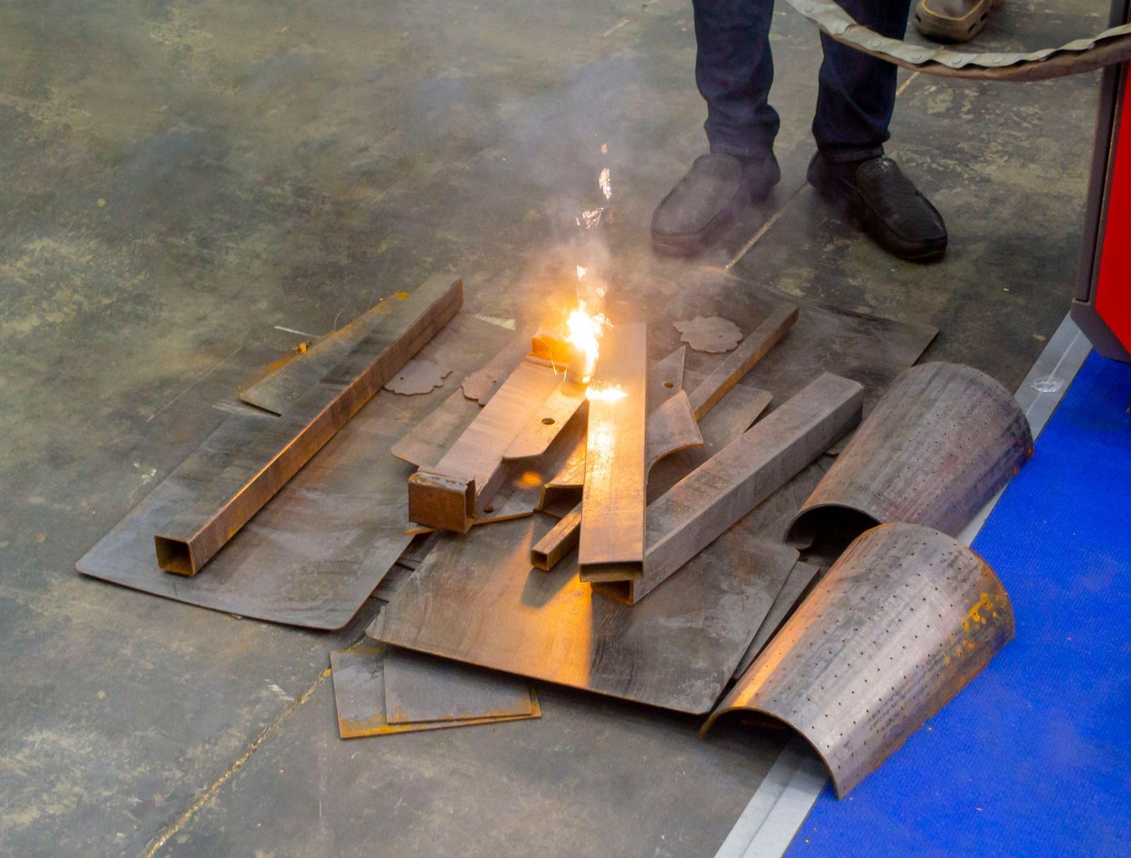 Person welding metal pieces on a sheet, sparks flying. Indoors, gray and blue floor.