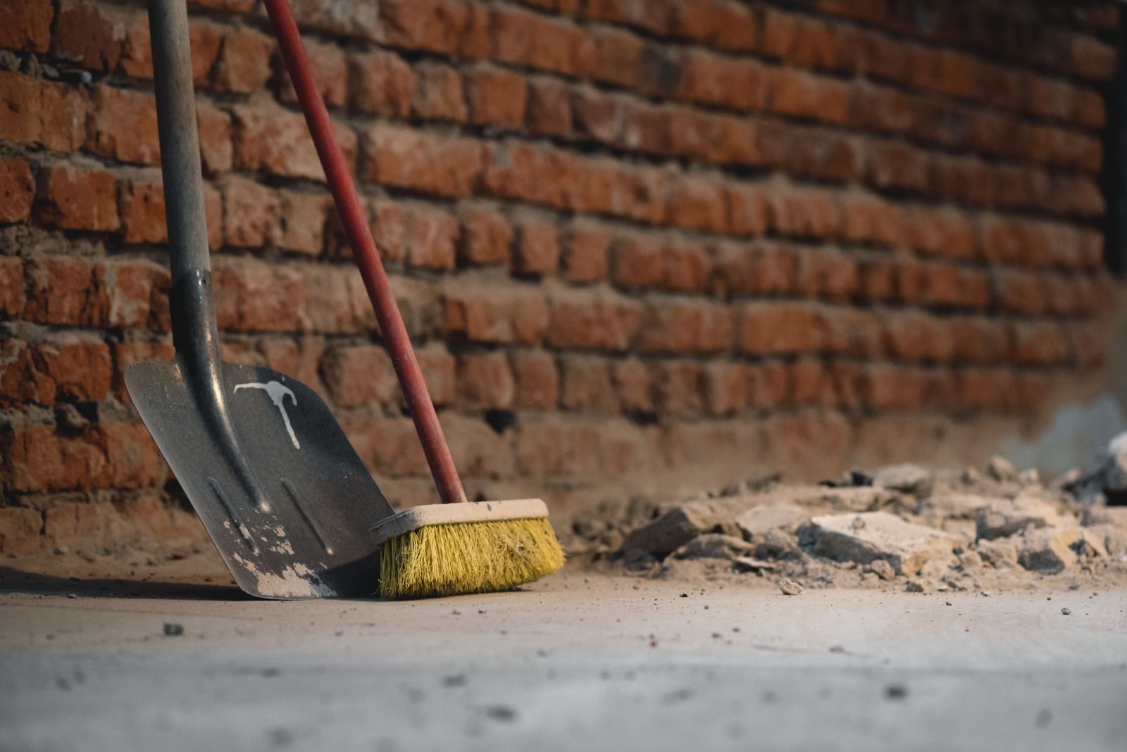 Shovel and broom resting against a brick wall, debris on ground, suggesting construction or cleanup.