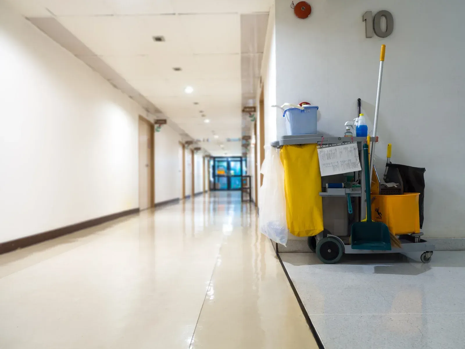 A janitorial cart with cleaning supplies sits in a hallway. White walls, shiny floor, and a distant blue door.