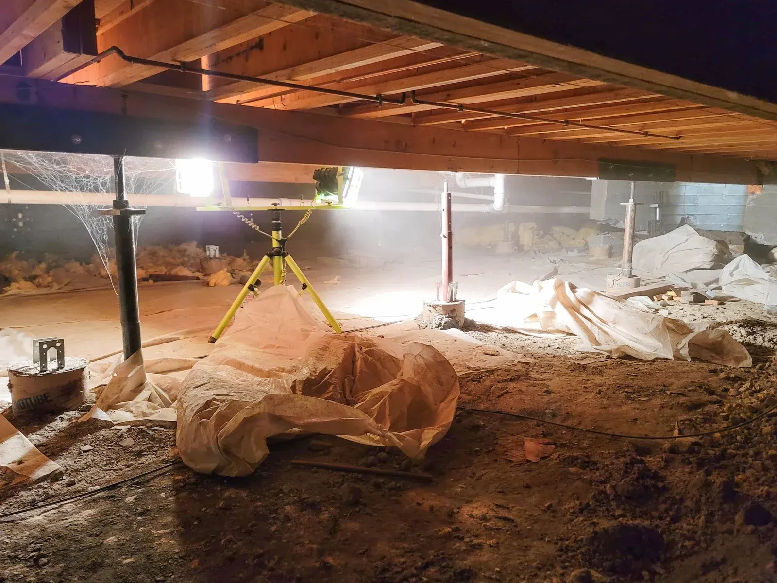 Underneath a structure, beams and jacks support it. A light illuminates the work area with stone debris.