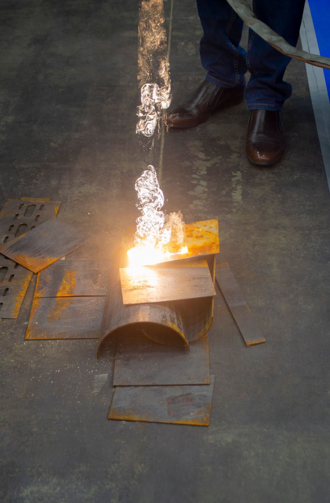 Person welding metal pieces on a concrete floor, sparks flying, with a blue line and rusty metal scraps nearby.