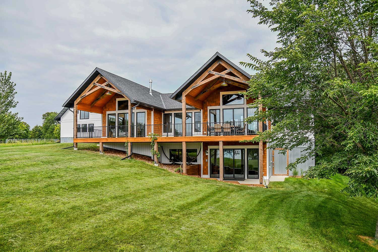 Exposed wood beams on porch house with glass windows and balconies on a grassy hillside under a cloudy sky.