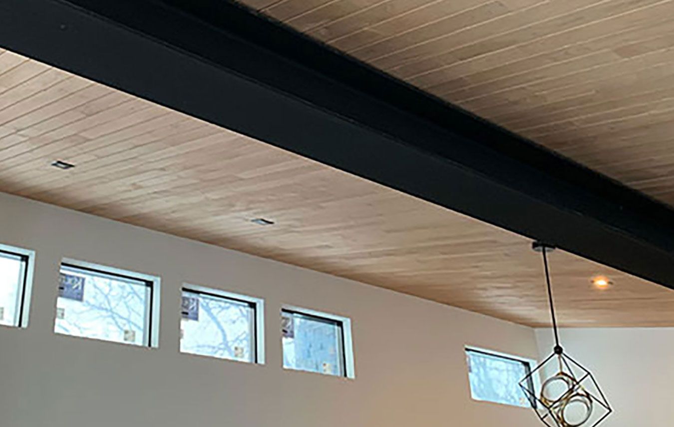 Interior view: wood-paneled ceiling, black beam, windows, pendant light, and beige walls.