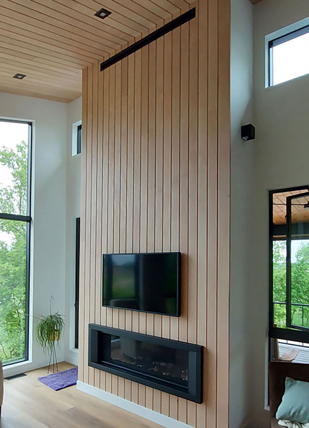 Wooden-paneled wall with a mounted TV and fireplace. Tall windows frame greenery. Light-colored wooden ceiling.