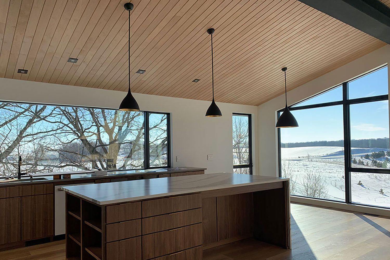 Modern kitchen with wood ceiling and island, large windows overlooking a snowy landscape.