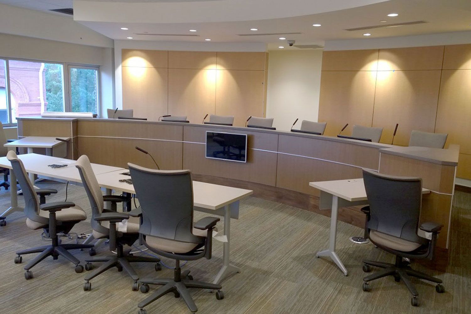 Conference room with a curved desk, chairs, and a monitor. Tables and chairs in the foreground, windows on the left.