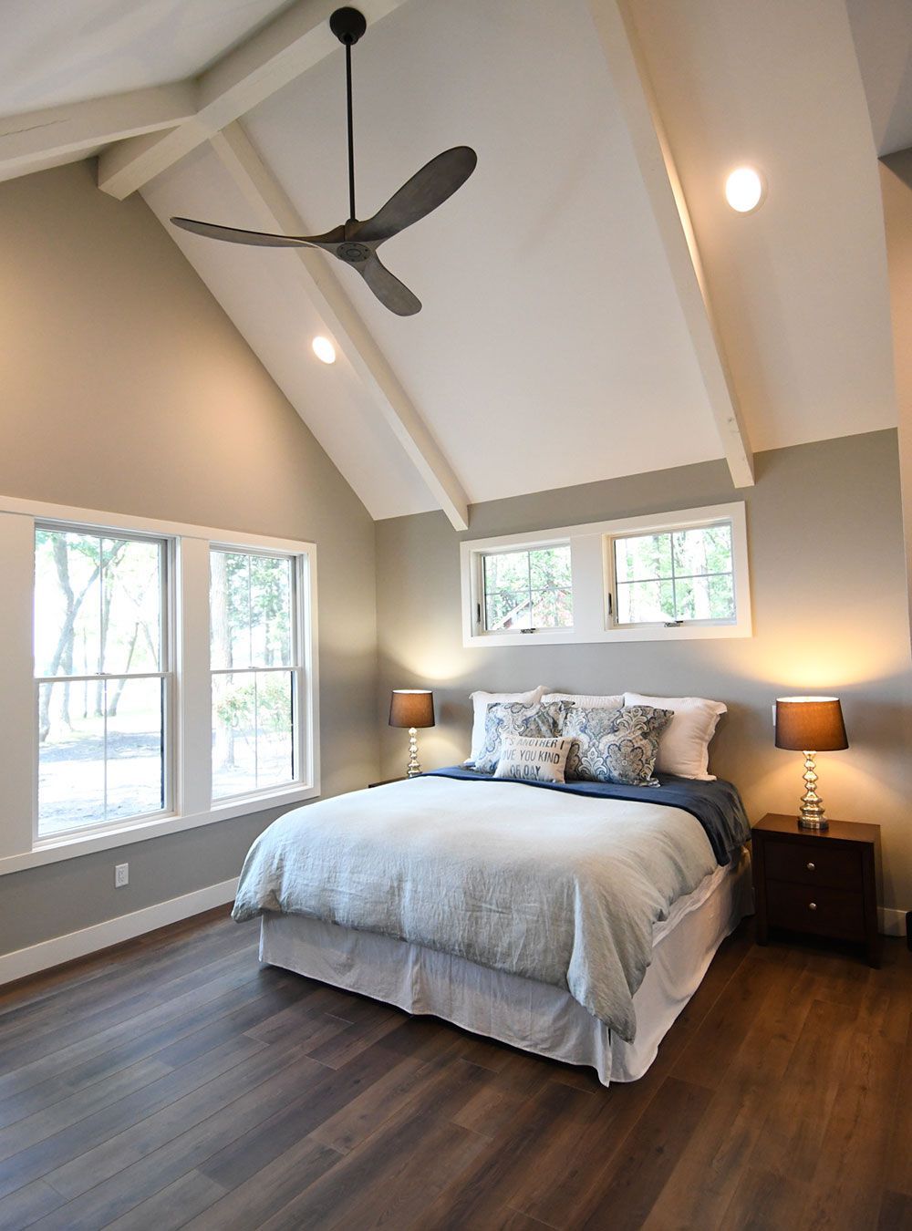 Bedroom with vaulted ceiling, bed with blue accents, dark wooden floors, windows, and a ceiling fan.